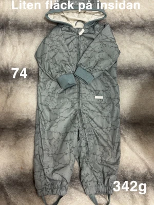 Blå/grön vindoverall med molnmönster - Säljer en blå/grön vindoverall med diskret molnmönster och huva. Overallen har dragkedja framtill, ribbade muddar vid ärmar och elastiska band vid benslut. Insidan är ljus och mjuk, perfekt för kyligare dagar.🌸Sparsamt använd. Lätt noppig på muddar. Något påverkade hällor. Lite missfärgad på på insidan kragen. Lite fläck på insidan. Färgen är blå/grön. Storlek 74 