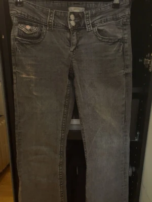 Grå tvättade flared jeans - Säljer ett par grå tvättade jeans med flare och klassisk femficksmodell. Jeansen har dubbla knappar i midjan och slitna detaljer framtill för en cool vintagekänsla. Perfekta för dig som gillar en avslappnad och trendig stil.