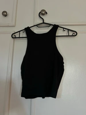 Svart croppad tanktopp - Snygg svart croppad tanktopp med hög halsringning och ärmlös design. Perfekt till jeans eller kjol för en clean och enkel look. Mjuk och stretchig känsla i materialet som sitter skönt på kroppen.
