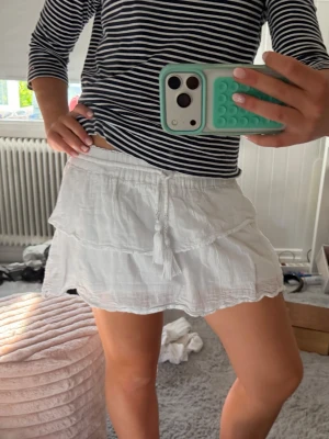 Vit volangkjol med snörning - Söt vit kortkjol med volanger och snörning i midjan. Kjolen har en luftig och lätt känsla, perfekt för varma dagar. Passar till både enkla toppar och mer dressade looks. Tillverkad i ett tunt, mjukt material. Från Gina