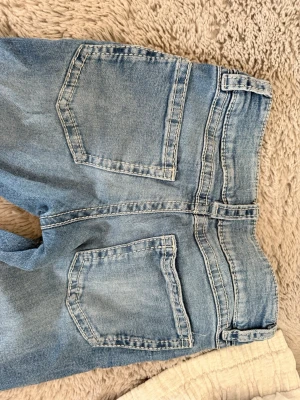 Ljusblå bootcut jeans med dubbla knappar - Säljer ett par ljusblå jeans med utsvängda ben och dubbla knappar i midjan. Jeansen har klassiska fem fickor och tydliga sömmar. Materialet är denim i bomull och passformen är avslappnad. Perfekta för en chill och trendig look. Från Gina tricot i storlek 158. De har en liten slitning nere på benet (se sista bilden).