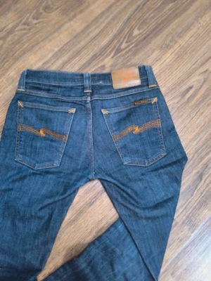 Mörkblå jeans från nudie  - Säljer ett par mörkblå jeans med raka ben och snygga kontrastsömmar i orange på bakfickorna. Jeansen har klassisk femficksdesign och läderpatch bak i midjan. Perfekta till vardagslooken och passar till det mesta i garderoben.