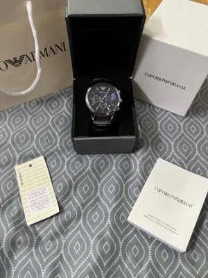 Emporio Armani silverfärgad klocka - Stilren klocka från Emporio Armani med silverfärgad länk i metall och mörk urtavla med flera små urtavlor. Klockan har klassisk rund form och tydlig logga vid tolvslaget. Kommer med originalask, manual och certificat.