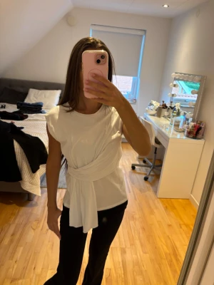 Vit omlottopp med knytband - Säljer en vit topp med omlottdetalj och breda knytband i midjan. Toppen är ärmlös och har en clean, modern look som passar perfekt till både jeans och kjol. Materialet känns mjukt och bekvämt mot huden.