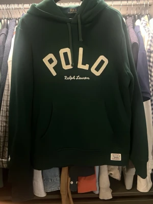 Grön hoodie från Polo Ralph Lauren - Mörkgrön hoodie med stor POLO-text i vitt framtill och Ralph Lauren-broderi under. Storlek M, hör av dig för fler bilder.