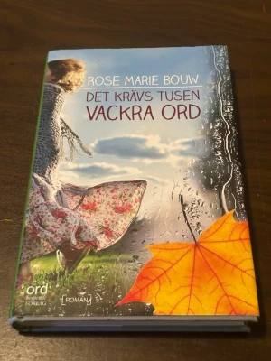 Det krävs tusen vackra ord - Romanen 'Det krävs tusen vackra ord' av Rose Marie Bouw har ett färgstarkt omslag med en flicka i blommig klänning, regndroppar på fönster och ett gult höstlöv. Boken ges ut av Ordberoende förlag och handlar om familj, känslor och livet på landet.