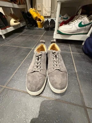 Gråa loubs - Snygga grå sneakers i mocka med vita sulor och röda detaljer undertill. Skorna har klassisk låg profil, rund tå och matchande grå skosnören. Insidan är ljusbeige för extra kontrast. Perfekta för en clean och stilren look.