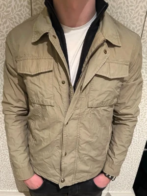 Massimo dutti overshirt - Stilren beige overshirt från massimo dutti med två stora bröstfickor och klassisk krage. Jackan har dragkedja och knappar framtill samt långa ärmar. Tillverkad i ett lätt material, perfekt för lager på lager under vår och höst.