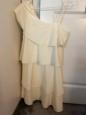 Gul oneshoulder klänning med volang - Säljer en elegant ljusgul oneshoulder klänning med volangdetaljer och lager på lager-design. Klänningen har ett axelband och är tillverkad i ett lätt, flödande material som passar perfekt till sommarens tillfällen.