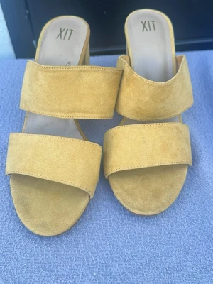 Gula slip-in sandaler från XIT - Snygga gula sandaler från XIT med två breda remmar i mockaimitation. Modellen är slip-in med öppen tå och låg klack, perfekt för en avslappnad och trendig look. Materialet ger en mjuk känsla och färgen sticker ut på ett coolt sätt.