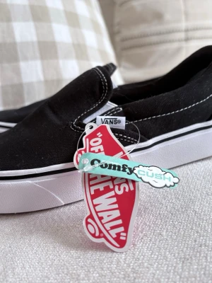 Svarta Vans Slip-On ComfyCush - Klassiska svarta Vans Slip-On sneakers med vit sula och kontrasterande vita sömmar. Modellen har ComfyCush-innersula för extra komfort och är tillverkad i canvasmaterial. Helt oanvända, köpta i Vansbutiken i Göteborg. 