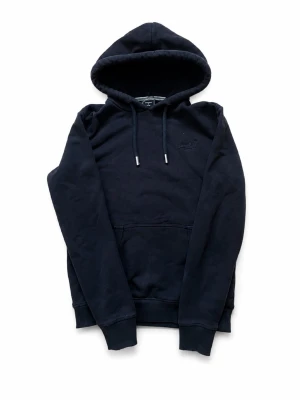 Svart hoodie från Superdry - Säljer en svart hoodie från Superdry med broderad logga på bröstet. Hoodien är i bra skick. Perfekt för en avslappnad och stilren look. Lätt att matcha med skön o varm.