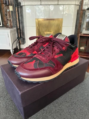 Helt Nya Valentino Garavani Rockrunners | Storlek 43 - Snygga sneakers från Valentino i vinrött, svart och rött med camoinspirerat mönster. Skorna har detaljer i mocka och läder, samt en beige sula och nitar på hälen. Box och tillbehör ingår! Nypris ca 5k! 