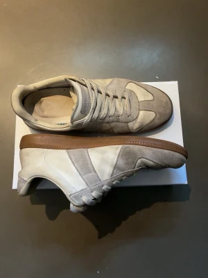 Maison margiela GATS - Snygga sneakers i beige och vitt med paneler i mocka och slät textil. Skorna har klassisk låg siluett, vita snören och en brun gummisula som ger en retrovibe. Perfekta för dig som gillar stilrena och tidlösa sneakers.