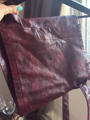 Vinröd shorts  i fejkläder - Säljer en stilren vinröd shorts i fejkläder med enkel design och en stor ytterficka. Väskan har justerbara axelband och en mjuk, något glansig yta. Perfekt för dig som vill ha en snygg och praktisk accessoar till skolan eller stan.
