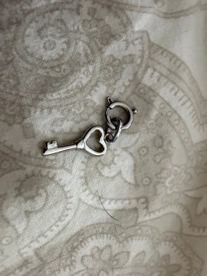Silverfärgad nyckelberlock - Söt berlock i form av en liten nyckel med hjärtformat handtag. Tillverkad i silverfärgad metall med en nyckel   med hänge till fästa i barlock armband/Halsband aldrig andvänt från Edblad 