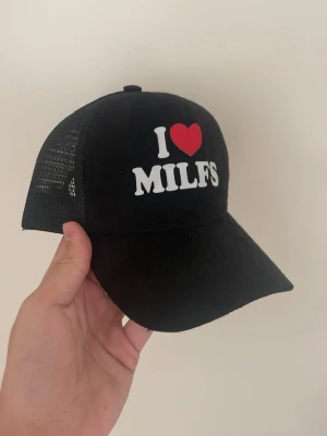 Svart truckerkeps med text - Säljer en svart truckerkeps med vit text och ett rött hjärta på framsidan där det står 'I ♥ MILFS'. Kepsen har böjd skärm och mesh på sidorna och bak, samt justerbar snapback. Perfekt för dig som vill sticka ut med din stil.