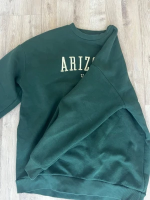 Mörkgrön sweatshirt Arizona USA - Mörkgrön sweatshirt med trycket 'ARIZONA U.S.A.' i vitt på bröstet. Tröjan har rund halsringning, ribbade muddar vid ärmslut och nederkant. Perfekt för en avslappnad och trendig look. Materialet ser ut att vara mjukt och bekvämt, troligen bomullsblandning💓