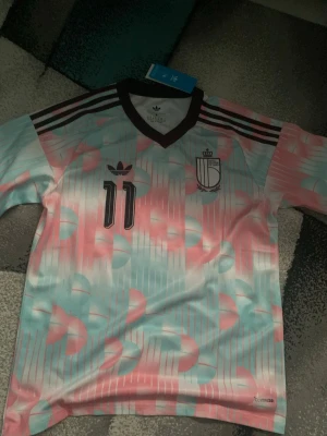 Belgiens landslagströja  - Säljer en retroinspirerad fotbollströja från Adidas i pastellfärger med rosa, ljusblått och vitt mönster. Tröjan har svarta detaljer, nummer 11 på bröstet och klassiska Adidas-ränder på axlarna. Tillverkad i lätt och ventilerande material.
