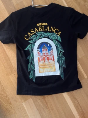 Svart t-shirt med Casablanca-tryck - Svart t-shirt från Casablanca med färgglatt tryck av en byggnad och texten 'Avenida Casablanca' både fram och bak. T-shirten har rund hals, korta ärmar och är gjord i mjuk bomull. Perfekt för dig som gillar grafiska prints och streetwear vibes.