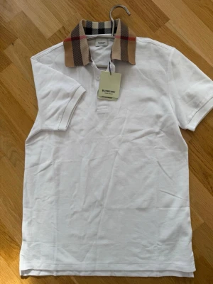 Vit Burberry t-shirt med rutigt krage - Snygg vit t-shirt från Burberry med klassisk kort ärm och ikonisk beige-rutig krage. T-shirten har knappar vid halsen och är tillverkad i bomull. Perfekt för dig som vill ha en stilren look med en touch av lyx.