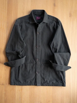 Shirtonomy Dark Green Chore Jacket, size L - Stilren svart overshirt med klassisk krage och knäppning framtill. Tillverkad i ett kraftigare bomullstyg som ger en avslappnad men ändå snygg look. Perfekt att bära över en t-shirt eller hoodie. Två diskreta fickor framtill och uppvikta ärmslut.