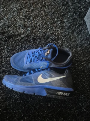 Nike Air Max blå sneakers - Säljer ett par blå Nike Air Max sneakers med vit swoosh och svart detalj vid hälen där det står 'Air Max'. Skorna har meshmaterial för bra ventilation och klassisk snörning. Perfekta för dig som gillar sportig stil och vill ha bekväma skor till vardags.