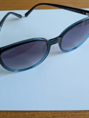 Blå ovala solglasögon från Swarovski - Snygga solglasögon från Swarovski med ovala bågar i transparent blå plast och mörka tonade linser. Perfekta för att ge din look en cool touch och skydda ögonen mot solen. Lätta och bekväma att bära hela dagen. Med fodral.