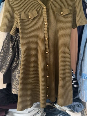 Olivgrön ribbad stickad klänning - Kortärmad olivgrön stickad klänning med ribbad struktur och dekorativa guldknappar längs hela framsidan. Två bröstfickor med lock och knappdetaljer ger en cool touch. Klänningen har en lätt utställd passform och v-ringad hals.