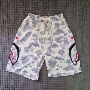 Camo shorts med hajmotiv och snörning - Säljer ett par loose-fit shorts med grått och vitt camouflagemönster, hajtänder- och ögonmotiv på sidorna samt snörning i midjan. Perfekta för dig som vill sticka ut med streetwear-vibe.