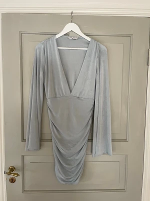 Ljusblå omlottklänning från NA-KD - Supersnygg ljusblå kortklänning från NA-KD med omlott-look, djup v-ringning och långa ärmar. Klänningen har draperad effekt framtill och är gjord i ett mjukt, glansigt material som känns lyxigt. Perfekt för dig som vill sticka ut med stil.