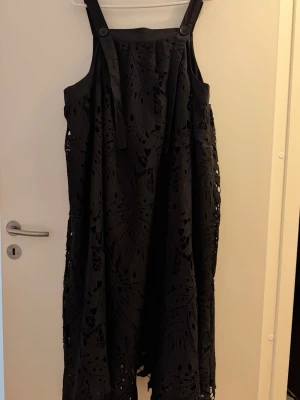 Svart klänning med spetsdetaljer - Säljer en svart klänning med breda axelband och vackra spetsdetaljer. Klänningen har ett utskuret mönster och en vågig kant nertill som ger en unik look. Perfekt för dig som vill sticka ut med en cool och elegant stil.