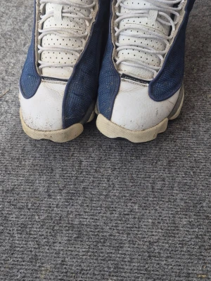 Air Jordan 13 - Säljer ett par höga sneakers i blått och vitt med perforerad ovandel och vadderad krage. Skorna har en unik paneldesign och synliga detaljer på hälen. Sulan är vit med beige inslag och ovandelen är i textil och syntet.