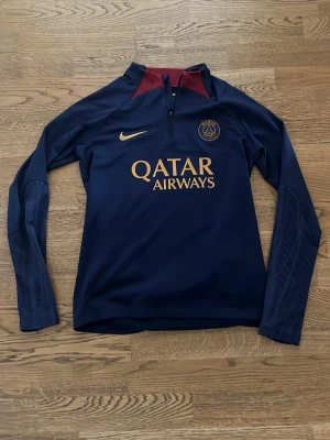 Mörkblå PSG träningströja Nike - Mörkblå långärmad träningströja från PSG med guldfärgade detaljer och Qatar Airways-tryck på bröstet. Halvlång dragkedja framtill och vinröda inslag vid kragen. Perfekt för fotbollsträning eller sportiga aktiviteter.