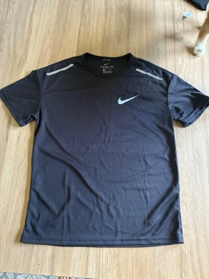 Svart Nike träningstopp - Snygg svart träningstopp från Nike med korta ärmar och rund hals. Toppen har diskreta grå detaljer på axlarna och den klassiska Nike-loggan på bröstet. Tillverkad i ett lätt och ventilerande syntetmaterial som passar perfekt för träning.
