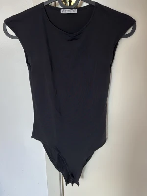 Svart body från ZARA - En stilren svart body från ZARA med rund halsringning och breda axlar. Tillverkad i ett mjukt och stretchigt material som sitter snyggt på kroppen. Perfekt att matcha med jeans eller kjol för en trendig look.