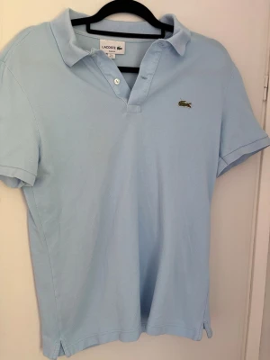 Ljusblå pikétröja från Lacoste - Klassisk ljusblå pikétröja från Lacoste med korta ärmar och den ikoniska krokodilloggan broderad på bröstet. Tröjan har krage och knappar framtill. Perfekt för en clean och sportig stil.