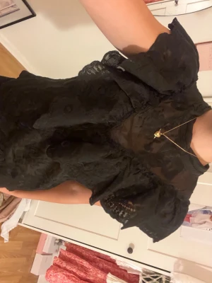 Svart blus med volang spets  - Säljer en svart blus med stora volanger och spetsdetaljer. Blusen är ärmlös och har ett transparent parti upptill som ger en cool look. Perfekt för dig som vill sticka ut med en unik topp med mycket detaljer.