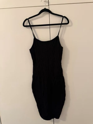 Svart kort klänning med struktur - Säljer en svart kort klänning med smala axelband och snygg struktur i tyget. Klänningen har en kroppsnära passform och är perfekt för dig som gillar minimalistisk stil med en twist. Materialet känns mjukt och stretchigt.