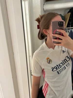 Vit Real Madrid matchtröja Adidas - Säljer en vit Real Madrid matchtröja från Adidas med klubbmärke och sponsortryck framtill. Tröjan har korta ärmar, V-ringning och rosa detaljer på sidorna. Perfekt för dig som älskar fotboll och vill representera ditt favoritlag.