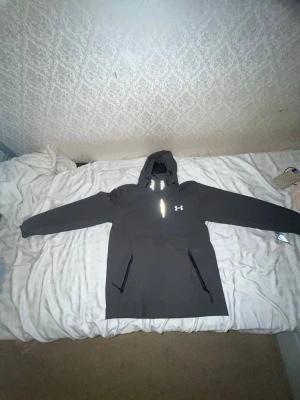 Svart vindjacka från Under Armour - Snygg svart vindjacka från Under Armour med huva och dragkedjor på fickorna. Jackan har en reflekterande logga på bröstet och är tillverkad i ett lätt, vindtätt material. Perfekt för blåsiga dagar och sportiga outfits.