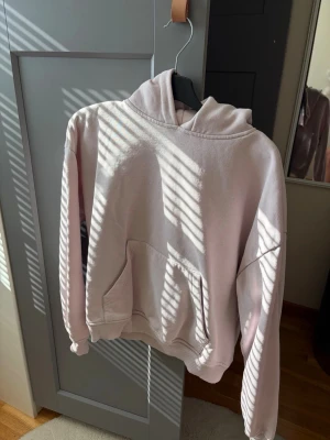 Ljusrosa hoodie med ficka - Säljer en ljusrosa hoodie med huva och stor magficka. Hoodien har ribbade muddar vid ärmslut och nederkant. Perfekt för en chill och avslappnad stil. Materialet känns mjukt och skönt mot huden.