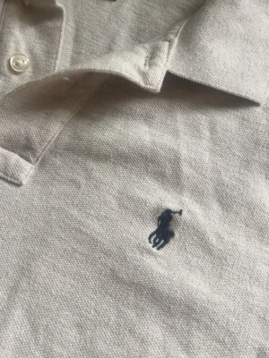 Beige pikétröja från Polo Ralph Lauren - Klassisk beige pikétröja från Polo Ralph Lauren med mörkblå broderad logga på bröstet. Tröjan har krage, korta ärmar och knappar vid halsen. Tillverkad i mjuk bomull som känns skön mot huden.