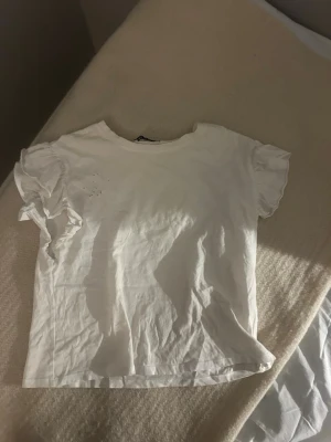 Vit t-shirt med volangärmar - Säljer en vit t-shirt med korta volangärmar. T-shirten har en enkel rund halsringning och är gjord i mjuk bomull. Perfekt för dig som gillar en clean och söt look med lite extra detalj på ärmarna. Den har ett litet märke uppe vid halsen typ men det är bara att toppen gick sönder av ett tvättmedel och använd endast en gång💓 