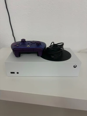 Xbox Series S med lila handkontroll - Säljer en vit Xbox Series S spelkonsol med en lila trådlös handkontroll och tillhörande laddkabel. Konsolen har en stilren, rektangulär design med svart cirkulärt ventilationsgaller på ovansidan. Perfekt för gaming och digitala spel.