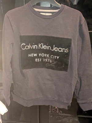 Mörkblå sweatshirt Calvin Klein Jeans - Mörkblå sweatshirt från Calvin Klein Jeans med stor svart patch och vit logga på bröstet. Rund halsringning och ribbade muddar vid ärmslut och nederkant. Inga synliga fläckar eller defekter. Skapa en bundle för bättre pris