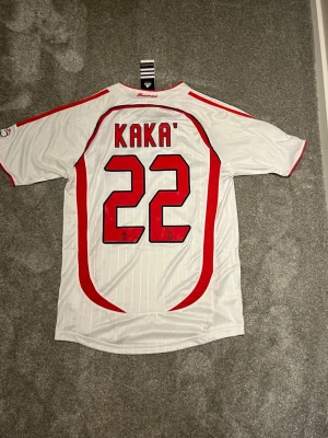 AC Milan Kaka' matchtröja vit - Säljer en vit AC Milan matchtröja från Adidas med Kaka' och nummer 22 på ryggen. Tröjan har röda och svarta detaljer, klubbmärke på bröstet och Champions League-finaltryck. Tillverkad i funktionsmaterial och har korta ärmar.