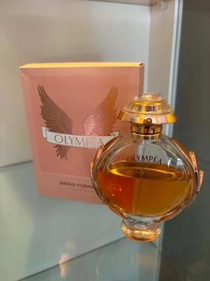 Olympea parfym från Paco Rabanne - Olympea parfym från Paco Rabanne i en elegant rund glasflaska med guldiga detaljer och en unik design inspirerad av antika troféer. Doften kommer i en rosa kartong med vingar och har en lyxig känsla. Perfekt för dig som gillar exklusiva parfymer.