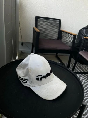 Vit keps från TaylorMade - Säljer en vit keps från TaylorMade med svart logga och text på framsidan samt TP5-tryck på sidan. Klassisk böjd skärm och justerbar passform. Perfekt för golf eller streetwear-stil. JAG VER ATT DEN ÄR EXTREMT SMUTSIG DÄRAV PRISET!
