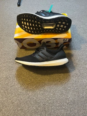 Svarta Adidas Ultraboost sneakers - Snygga svarta Adidas Ultraboost sneakers med stickad ovandel och vita Boost-sulor. Skorna har klassiska tre ränder på sidan och svart yttersula med grepp. Perfekta för dig som vill ha både stil och komfort i vardagen.
